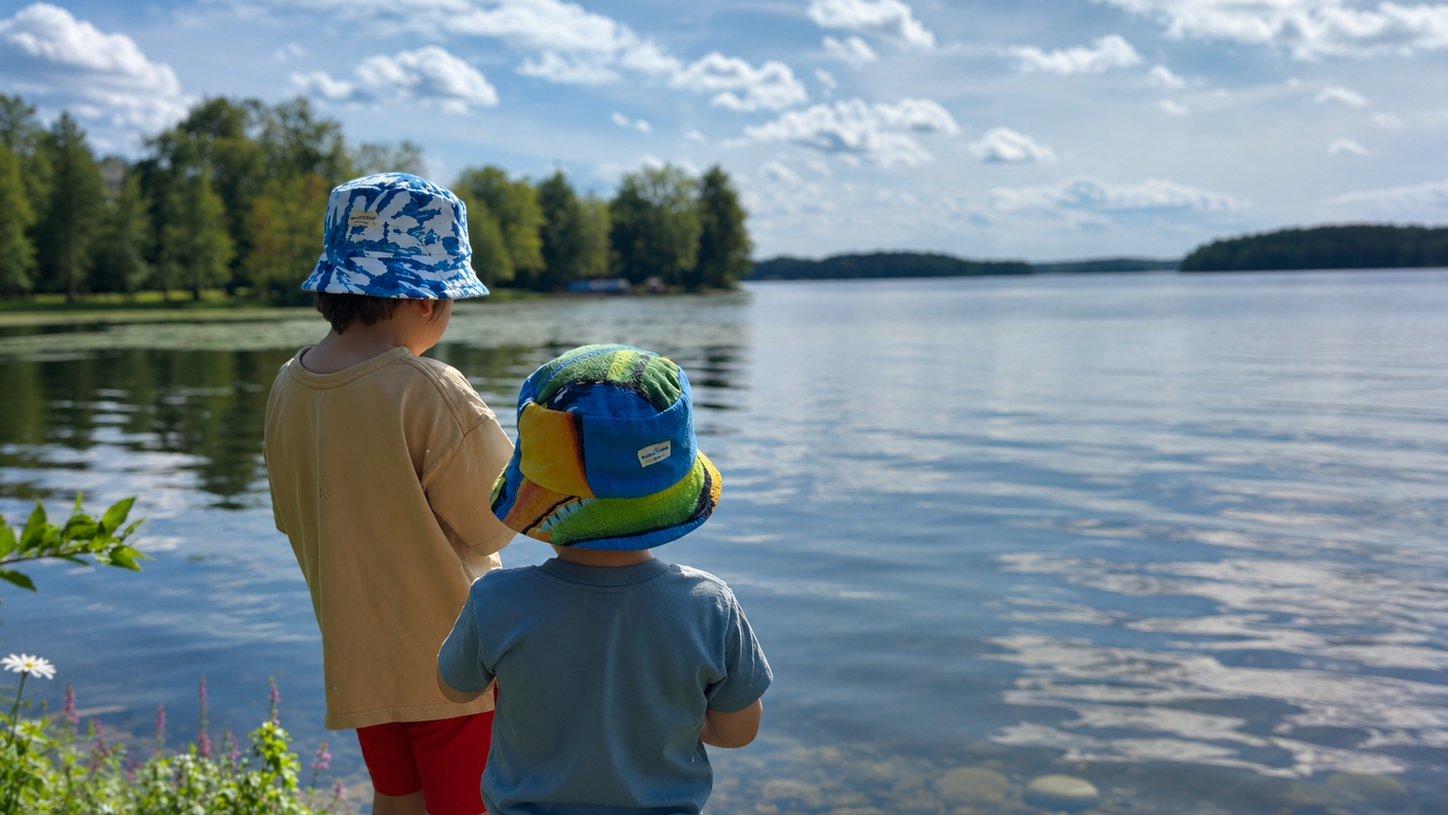 Two children standing on a lakeshore, seen from behind, wearing colorful patterned bucket hats and casual summer clothes while looking out over the water under a partly cloudy sky.